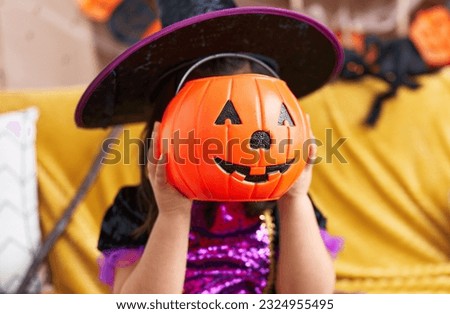 Adorable hispanic girl having halloween party holding pumpkin basket over face at home