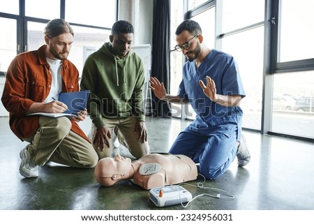 young paramedic explaining cardiac resuscitation techniques to interracial participants near CPR manikin with automated external defibrillator, effective life-saving skills and techniques concept Royalty-Free Stock Photo #2324956031