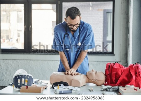young paramedic in blue uniform and eyeglasses practicing chest compressions on CPR manikin near defibrillator and first aid kit during medical seminar, life-saving skills development concept Royalty-Free Stock Photo #2324956063