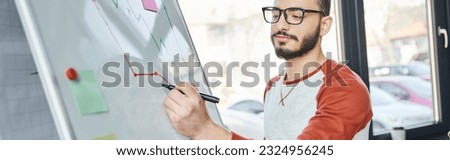 young bearded businessman in eyeglasses drawing infographics with felt pen on flip chart in contemporary office, working on startup project, creative thinking, entrepreneurship, banner