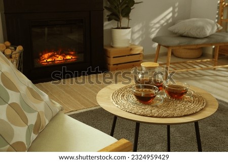 Teapot and cups of drink on coffee table near stylish fireplace in cosy living room. Interior design