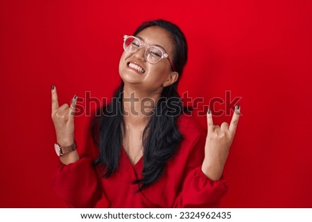 Asian young woman standing over red background shouting with crazy expression doing rock symbol with hands up. music star. heavy music concept. 