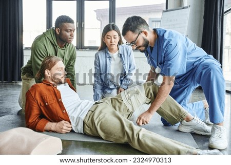 first aid seminar, medical instructor in eyeglasses and uniform applying compression bandage on leg of man near asian woman and african american, importance of emergency preparedness concept Royalty-Free Stock Photo #2324970313