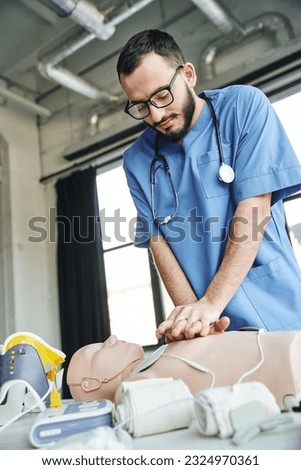 bearded professional paramedic in eyeglasses and blue uniform practicing chest compressions on CPR manikin near defibrillator and compressive bandages, critical skills development concept Royalty-Free Stock Photo #2324970361