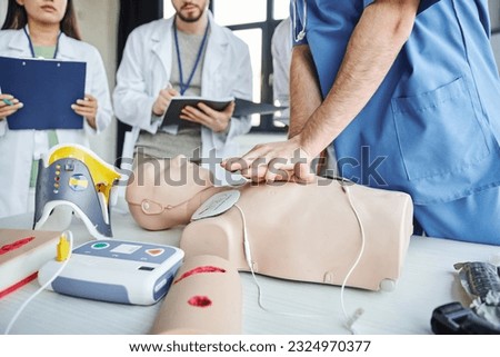 partial view of paramedic practicing chest compressions on CPR manikin near defibrillator, neck brace and students with clipboard and notebook, life-saving skills hands-on learning concept Royalty-Free Stock Photo #2324970377