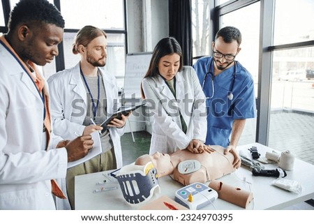 young asian woman in white coat doing chest compressions on CPR manikin near instructor, medical equipment and multicultural students with notebooks, emergency situations response concept Royalty-Free Stock Photo #2324970395