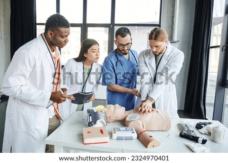 multiethnic students with notebooks looking at man doing chest compressions on CPR manikin near healthcare worker and medical equipment in training room, emergency situations response concept Royalty-Free Stock Photo #2324970401