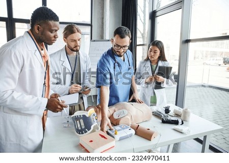 healthcare worker showing defibrillator, CPR manikin and medical equipment to young multiethnic group in white coats standing with notebooks in training room, emergency situations response concept Royalty-Free Stock Photo #2324970411