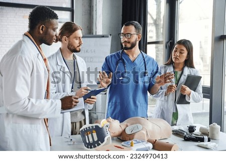 multicultural students in white coats writing near healthcare worker gesturing and talking next to CPR manikin and medical equipment on first aid seminar, emergency situations response concept Royalty-Free Stock Photo #2324970413