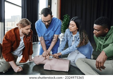first aid training, medical instructor showing cardiopulmonary resuscitation on CPR manikin near multiethnic participants in training room, effective life-saving skills and techniques concept Royalty-Free Stock Photo #2324975365