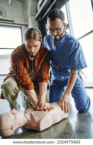 first aid hands-on learning, professional paramedic assisting young man practicing chest compressions on CPR manikin, effective life-saving skills and emergency preparedness concept Royalty-Free Stock Photo #2324975403