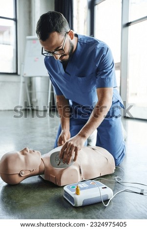 first aid training seminar, young medical instructor in uniform and eyeglasses applying defibrillator pads on CPR manikin, cardiac resuscitation, health care and life-saving techniques concept Royalty-Free Stock Photo #2324975405