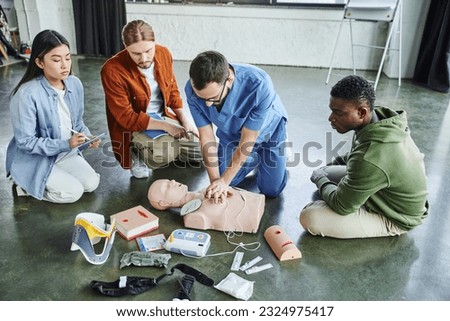 first aid seminar, multiethnic team looking at medical instructor doing chest compressions on CPR manikin near defibrillator, wound care simulators, neck brace, bandages and compression tourniquets Royalty-Free Stock Photo #2324975417