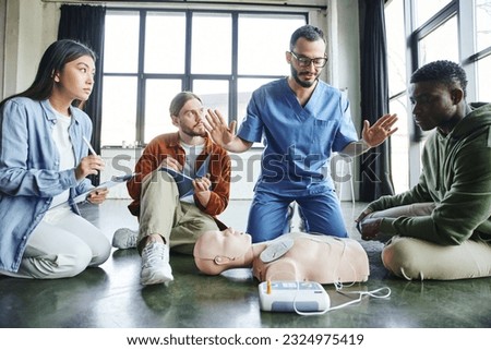 professional paramedic gesturing and talking to multiethnic participants near CPR manikin and defibrillator during first aid seminar in training room, health care and life-saving techniques concept Royalty-Free Stock Photo #2324975419