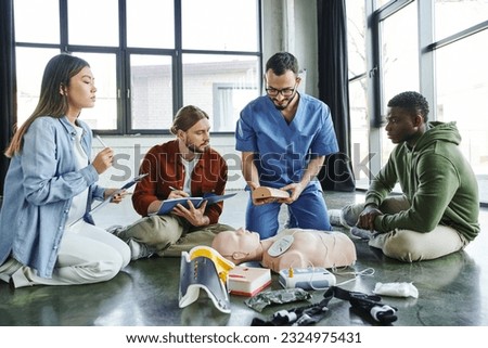 medical instructor showing wound care simulator to multiethnic team with clipboards near CPR manikin and medical equipment in training room, effective life-saving skills concept Royalty-Free Stock Photo #2324975431