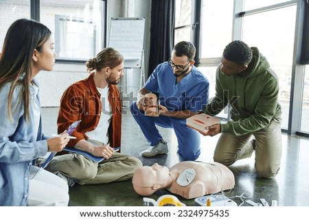medical instructor and african american man holding wound care simulators near CPR manikin, defibrillator and multiethnic participants with notebook and clipboard, life-saving skills concept Royalty-Free Stock Photo #2324975435