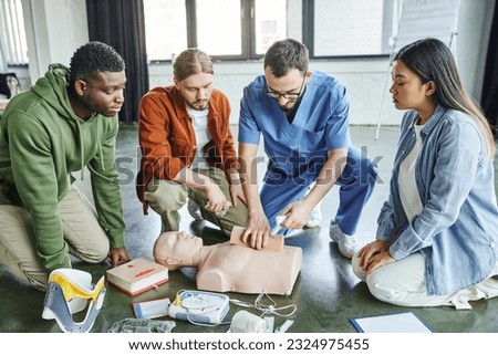 healthcare worker tamponing wound on simulator with bandage while showing life-saving skills to multicultural team near CPR manikin, defibrillator and medical equipment, emergency response concept Royalty-Free Stock Photo #2324975455