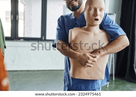 first aid training seminar, partial view of bearded medical instructor in uniform practicing life-saving techniques in case of chocking on CPR manikin, emergency situations preparedness concept Royalty-Free Stock Photo #2324975461