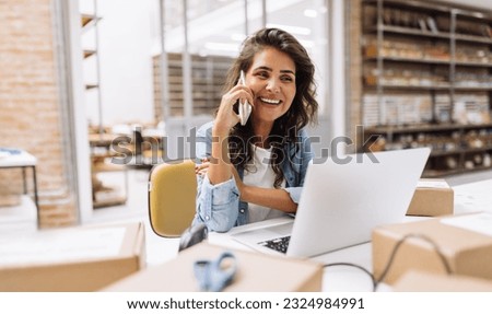 Happy young businesswoman speaking on the phone while working in a warehouse. Online store owner making plans for product shipping. Creative female entrepreneur running an e-commerce small business. Royalty-Free Stock Photo #2324984991