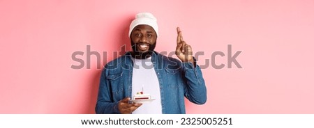 Handsome african-american guy celebrating birthday, making wish with fingers crossed, holding bday cake with candle, standing against pink background.