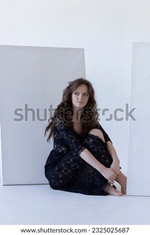 Isolated Young Woman Sits Near White Blank Large Stretched Canvas for Painting. Pretty Female Artist Poses With Empty Boards On White Background Indoor. Mockup. Vertical Plane. High quality photo