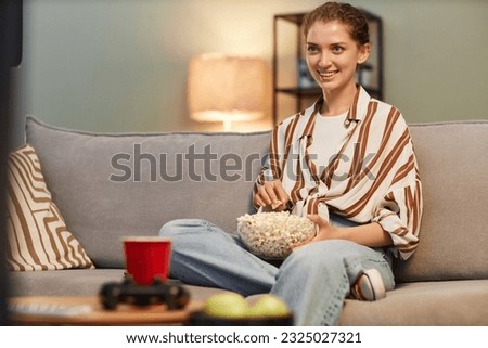 Portrait of smiling young woman watching TV at home and eating popcorn, copy space Royalty-Free Stock Photo #2325027321