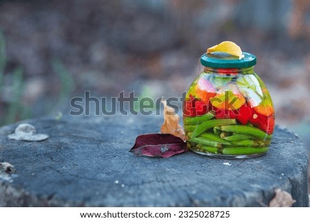 
A jar of canned red and green chili peppers in apple cider vinegar on an oak stump in a garden with fallen yellow and red leaves