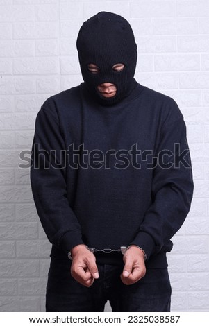 Man with black mask and outfit suspect of a robbery, wearing handcuffs in front of white background