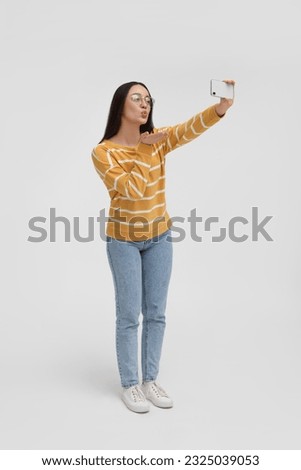 Young woman taking selfie with smartphone and blowing kiss on white background