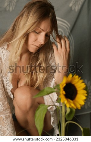 beautiful attractive blonde woman sadly looking down, sitting on the ground, bohemian background, wearing crocheted dress, kneeling in front of sunflower
