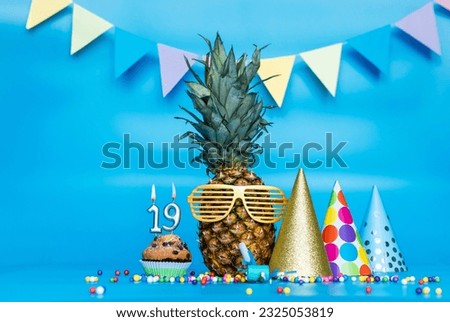 Creative background with pineapple character in sunglasses copy space. Happy birthday background with muffin or cake with candle number  19. Anniversary holiday decorations on a blue background.