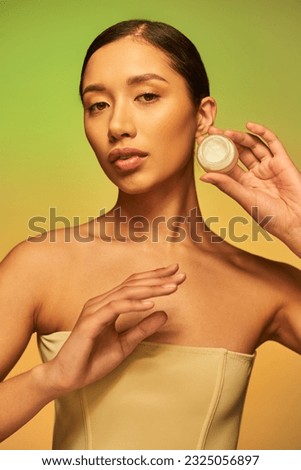 beauty photography, young asian woman with bare shoulders holding cosmetic jar with cream on green background, brunette hair, beauty industry, glowing skin, skin care concept