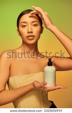 product presentation, young asian woman with bare shoulders holding cosmetic bottle with beauty product and looking at camera on green background, brunette hair, glowing skin