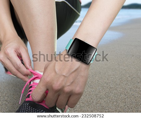 female hand tying shoelaces wearing smartwatch with bright blue watchband blank black glass bent touchscreen on natural sea beach background