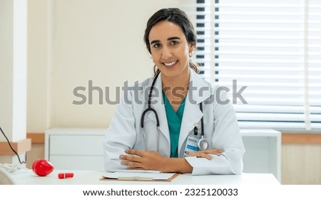 Woman female doctor portrait in white coat gown with stethoscope sit and smile to front. Medical student attractive friendly wear white coat smiling and work on table in patient room