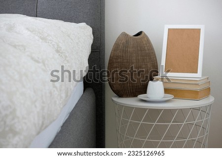 Brown ceramic vase, white cup of coffee with saucer and white frame mock up standing on old books on white bedside table. Beige floral bedding and gray upholstered bed in bedroom. Neutral interior.