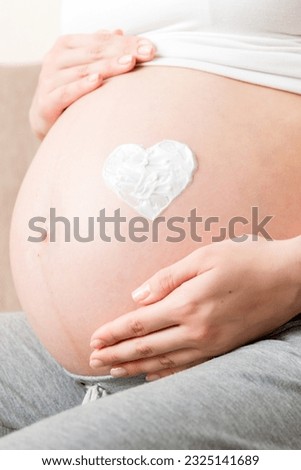 a pregnant girl sits at home on the bed and smears heart an anti-stretch mark cream on her stomach. Pregnancy, motherhood, preparation and expectation concept.