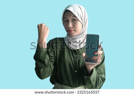 A portrait of a happy Asian Muslim woman wearing a headscarf and green shirt showing her phone screen, isolated on blue background