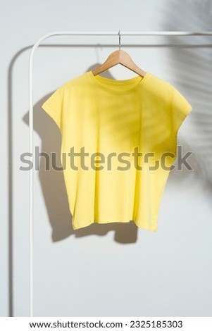 Blank yellow t-shirt mockup, template on wooden hanger displayed on a wite clothes rack. White background with palm leaf shadow.