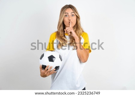 Surprised Young beautiful woman holding football ball over white background makes silence gesture, keeps finger over lips and looks mysterious at camera