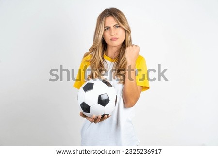 Young beautiful woman holding football ball over white background shows fist has annoyed face expression going to revenge or threaten someone makes serious look. I will show you who is boss