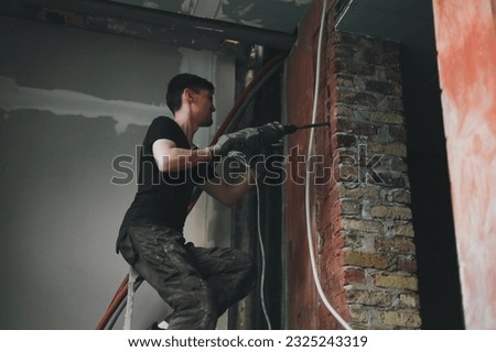 A young Caucasian man recognizable in a black uniform emotionally breaks a hole in a brick wall with a puncher with a large screwdriver while sitting on a stepladder,close-up bottom view with selectiv