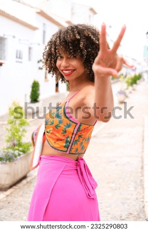 A girl with afro hair and dressed in bright colors makes the victory sign with her hand while smiling.