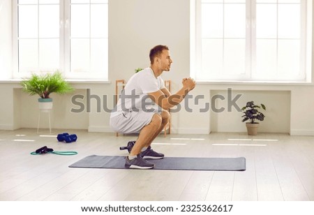 Side view of a fit, sporty, young man in activewear having a sports workout at the gym or at home, standing on a fitness workout mat and doing a squatting exercise for his leg and glute muscles Royalty-Free Stock Photo #2325362617