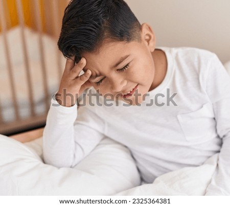 Adorable hispanic boy sitting on bed thinking at bedroom Royalty-Free Stock Photo #2325364381