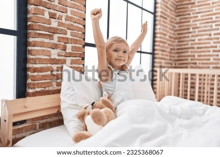 Adorable blonde girl sitting on bed with winner expression at bedroom