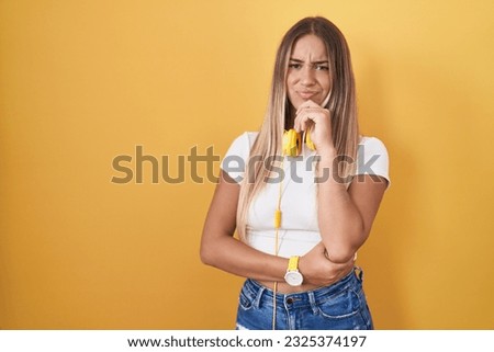Young blonde woman standing over yellow background wearing headphones looking confident at the camera smiling with crossed arms and hand raised on chin. thinking positive. 