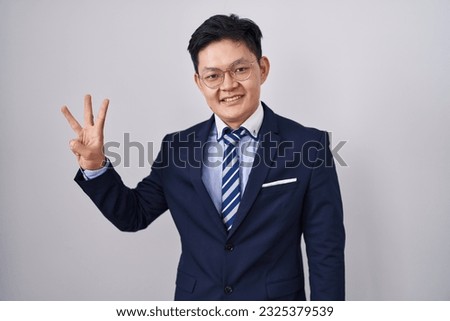 Young asian man wearing business suit and tie showing and pointing up with fingers number three while smiling confident and happy. 