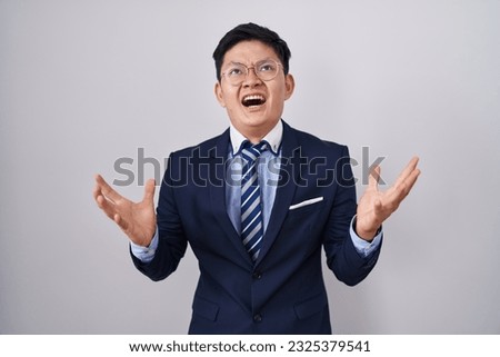Young asian man wearing business suit and tie crazy and mad shouting and yelling with aggressive expression and arms raised. frustration concept.  Royalty-Free Stock Photo #2325379541