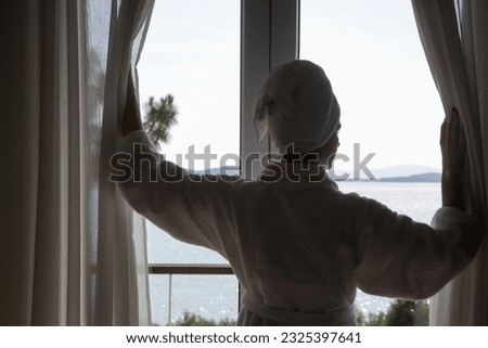 Woman in a bathrobe opening curtains in room in the morning with beautiful peaceful seaview Royalty-Free Stock Photo #2325397641